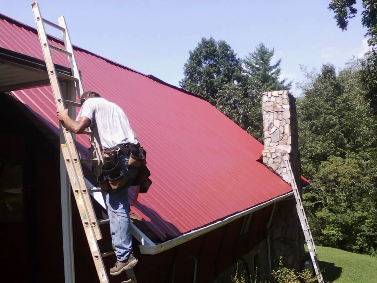 Roofer on a ladder beside a red metal roof and stone chimney — Soffit & Fascia Repair by Roofing Brothers in Leawood